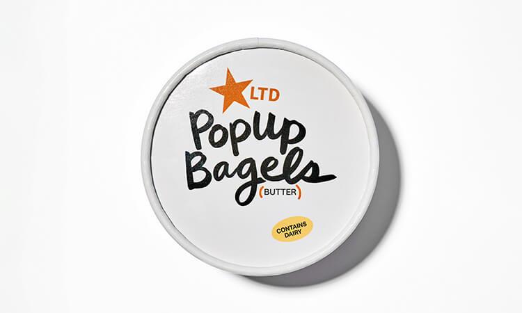 Lid of a butter container labeled "Popup Bagels LTD" with an orange star and "Contains Dairy" note on a white background.