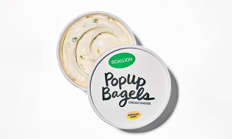 Cream cheese container labeled "Popup Bagels" with "Scallion" flavor. Lid partially open, revealing creamy cheese with visible scallion bits.